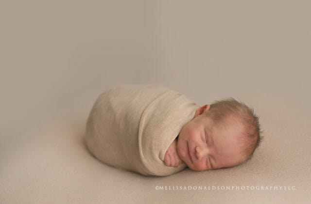 New baby wrapped in a beige stretchy swaddle wrap, lying on his side smiling. 