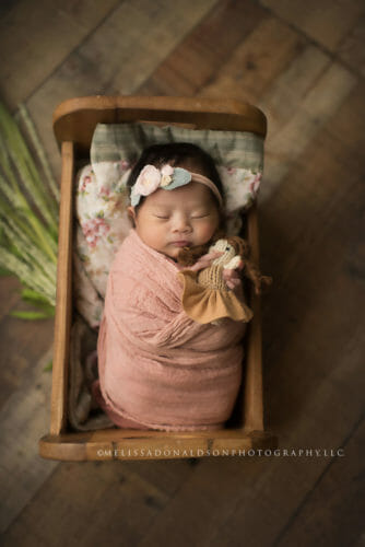 photo of a baby girl in a newborn prop bed, holding a baby doll and wearing  a pink swaddled wrap