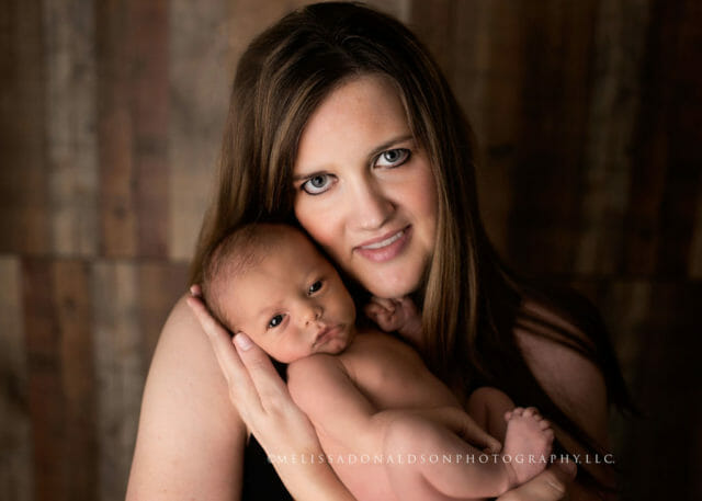 Photograph of new mom wearing a black tank top holding her seven day old baby with a wood backdrop
